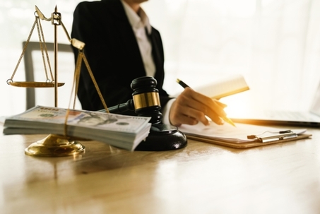 A professional is seated at a desk, with a gavel and a stack of money in the foreground, while writing in a notebook next to a laptop.